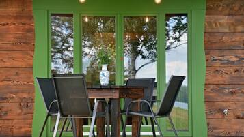 Una cabaña de madera con puerta y ventanas verdes, una mesa de comedor con sillas, y un florero con flores en el alféizar.