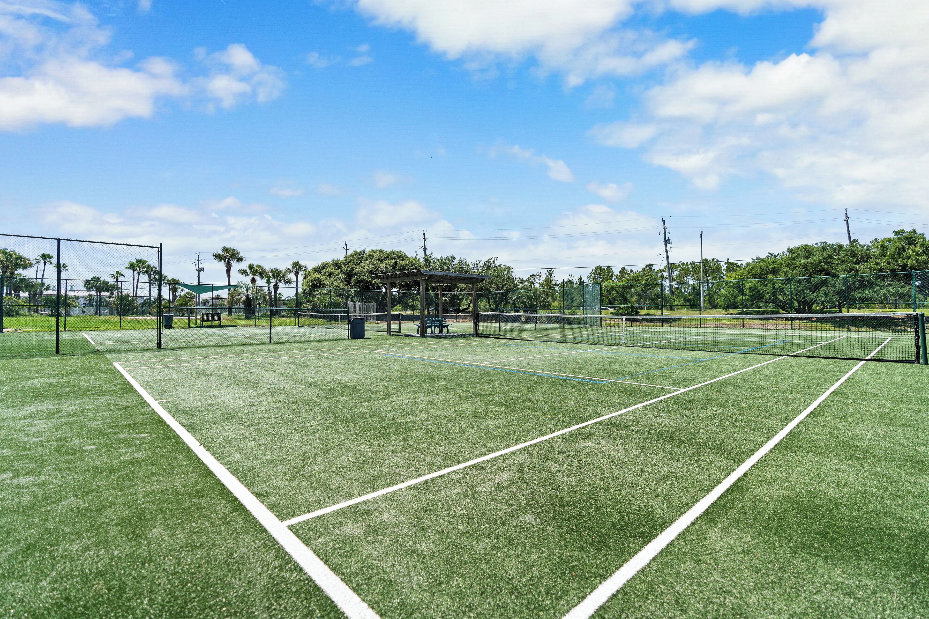 A tennis court with a net, surrounded by a fence, trees, and a gazebo.