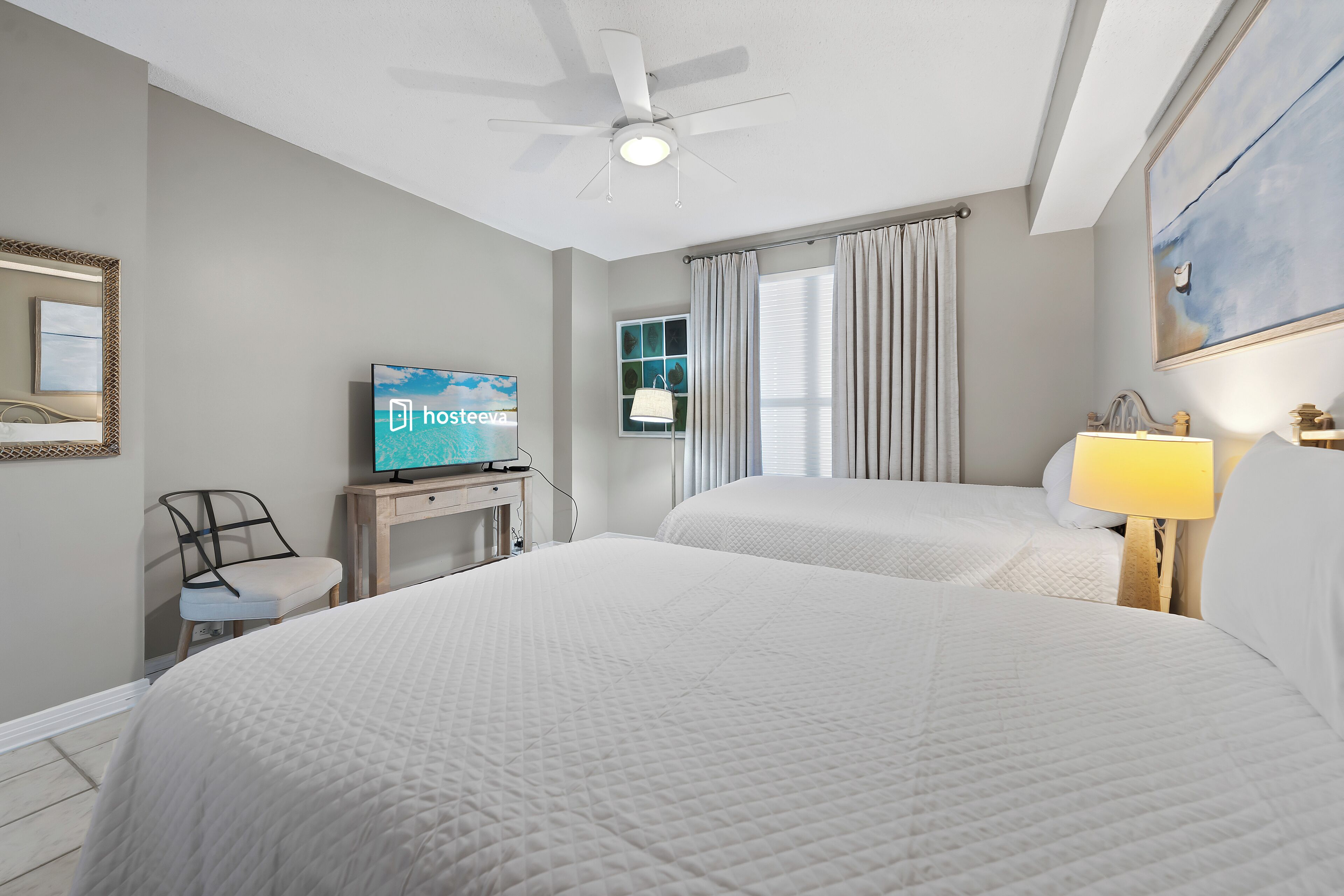 A hotel room with a large bed, a desk with a television, a ceiling fan, and a window with curtains.