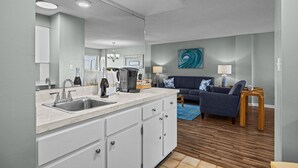 Una cocina moderna con encimera de mármol y armarios blancos, un comedor con sofá azul y mesita de madera, y una sala de estar con alfombra azul y un cuadro en la pared.
