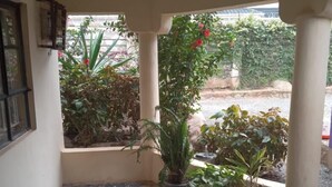 Un porche cubierto con suelo de baldosas, plantas en macetas y una ventana con vista a un jardín.