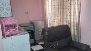 Una sala de estar con piso a cuadros, un sillón de cuero marrón, una silla rosa, un refrigerador y una ventana con cortinas estampadas.