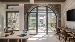 A modern interior with a large arched doorway leading to a pool area, a round table with a laptop, and several chairs. - Elounda Riviera Villa (Elounda)