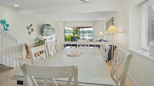 A dining area with a glass table, white chairs, a chandelier, and a view of greenery outside. - Waterfront Sea Pines Villa | 3 Private Suites, Balconies, Dock Access (Hilton Head Island)