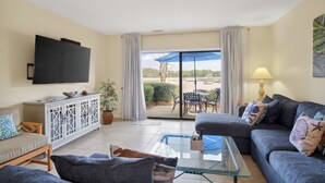 A living room with a sofa, a glass coffee table, a TV, a cabinet, and a view of the outdoors through a sliding glass door. - Waterfront Sea Pines Villa | 3 Private Suites, Balconies, Dock Access (Hilton Head Island)