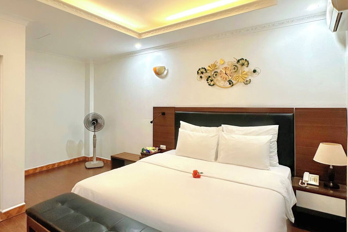 Una habitación de hotel con una cama grande, una mesita de noche con lámpara, un reloj de pared y un ventilador de techo.
