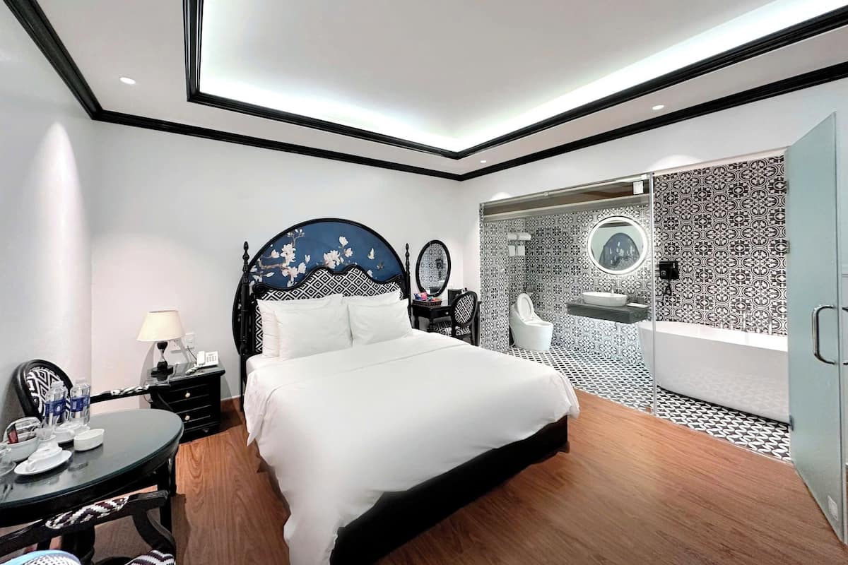 Una habitación de hotel moderna con una cama grande, un escritorio con lámpara, una silla y un baño con lavamanos y espejo.
