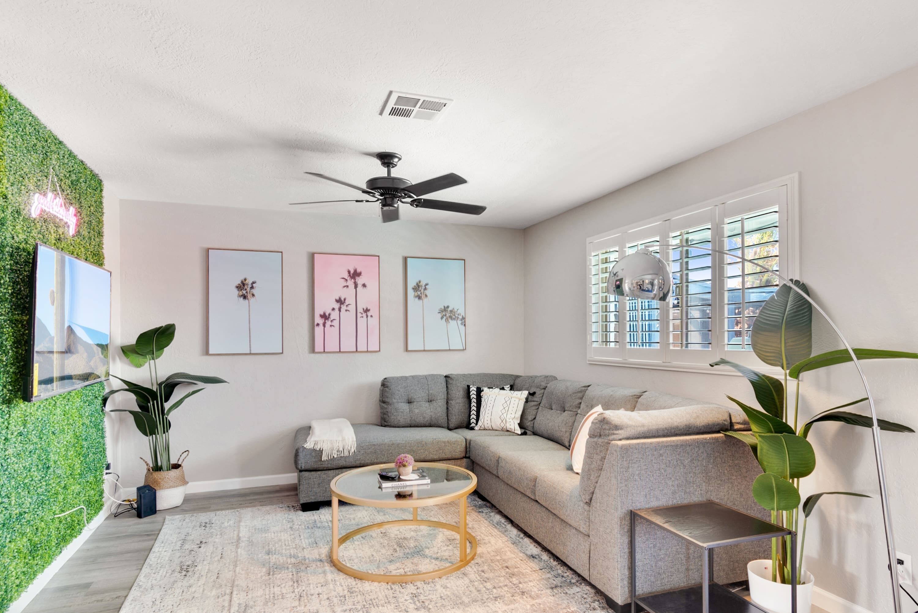 A modern living room with a grey sectional sofa, a round coffee table, and a ceiling fan. There are framed artworks on the wall and a large window with blinds.