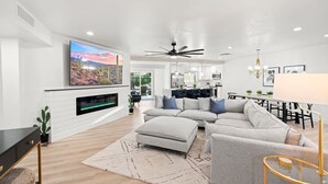 A modern living room with a sectional sofa, a flat-screen TV mounted on the wall, a ceiling fan, and a dining area with a chandelier. - Scottsdale Classic - Luxury Home w/ 5 Beds & Pool! (Scottsdale)