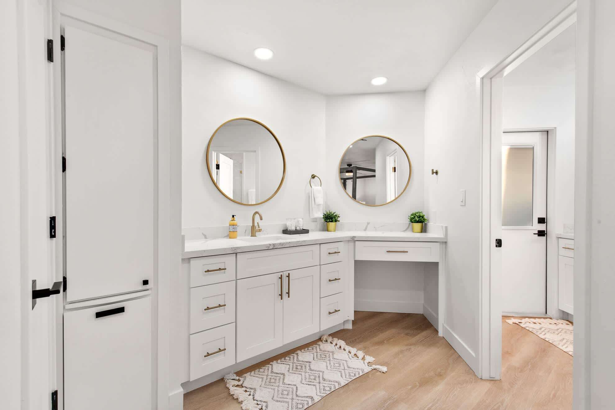 Une salle de bain moderne avec des meubles blancs, deux miroirs ronds dotés de cadres dorés, un plan de toilette blanc et un tapis à motifs sur le sol.
