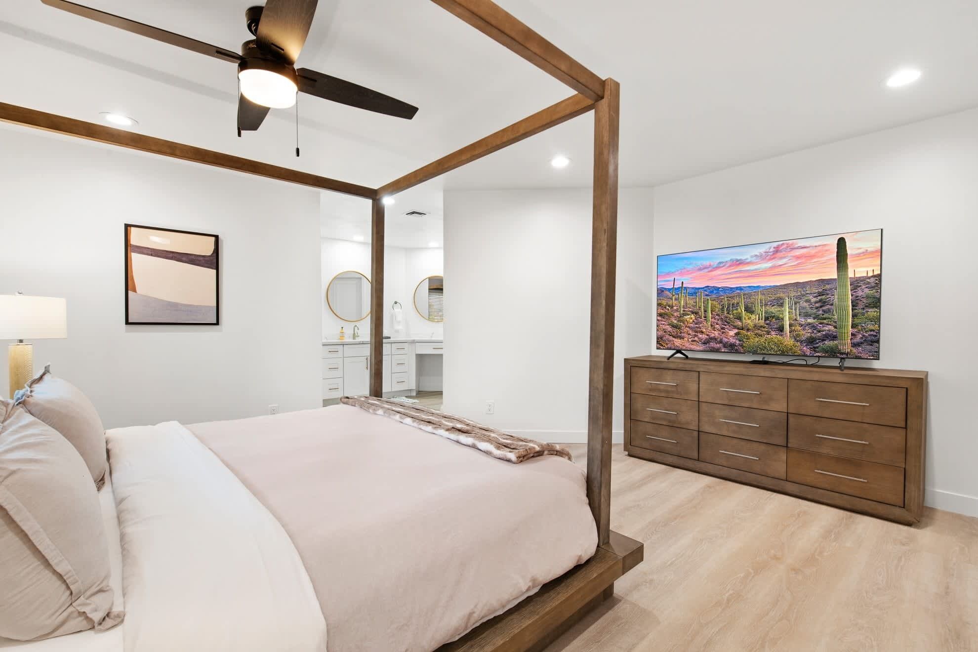 A bedroom with a four-poster bed, a ceiling fan, a large flat-screen TV, a dresser, and a framed picture on the wall.