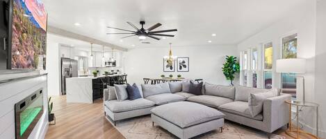 A modern living room with a sectional sofa, a ceiling fan, and a kitchen in the background.