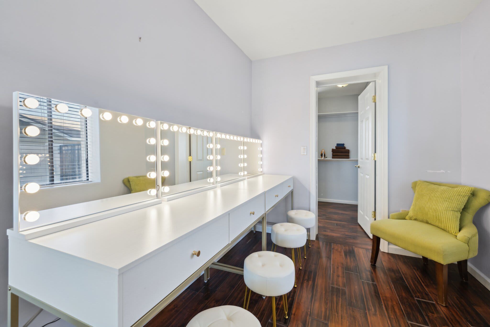 A bathroom with a vanity, mirror with lights, stools, and a chair.