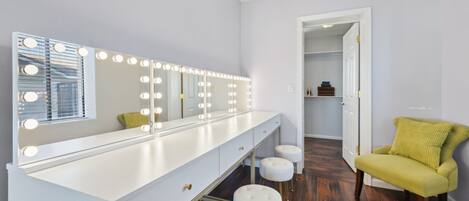 A bathroom with a vanity, mirror with lights, stools, and a chair.