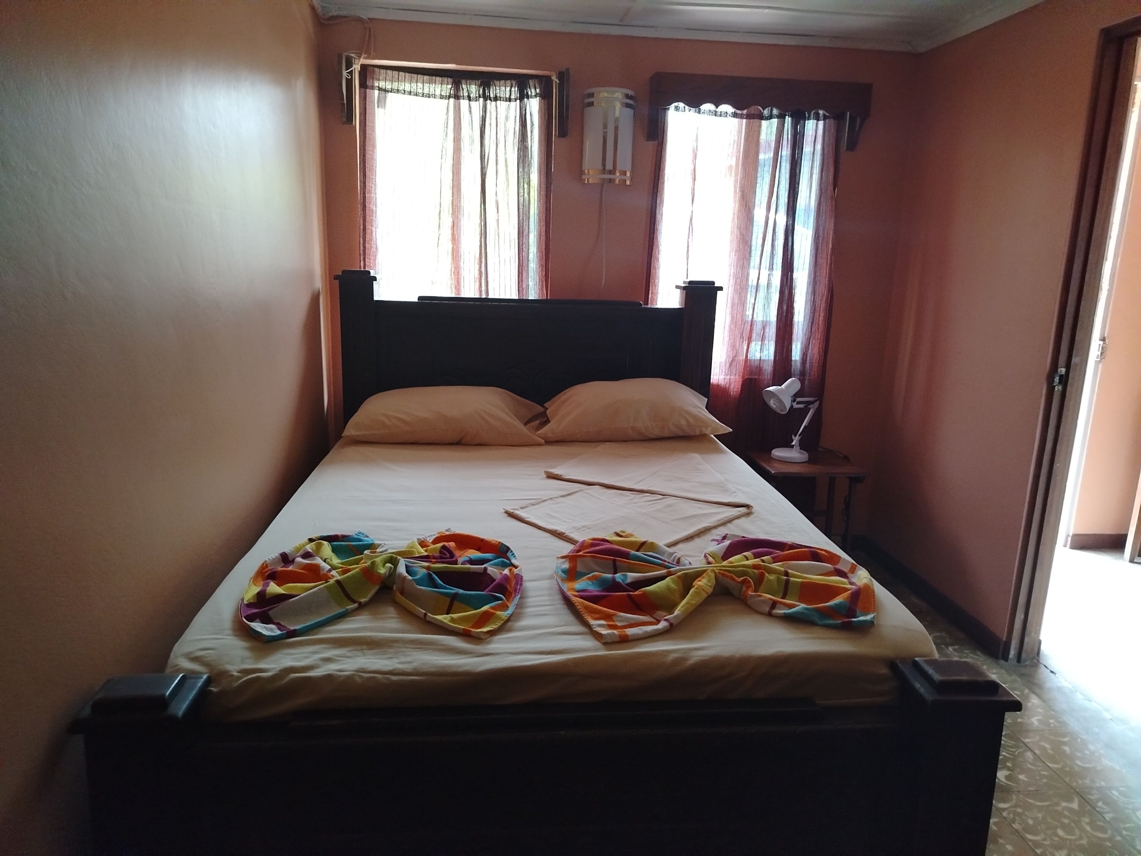 A single bed with a plain mattress, two pillows, a bedside table with a lamp, and a window with curtains.