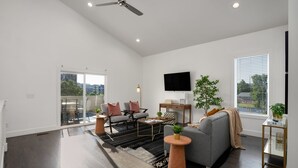 A modern living room with a grey sofa, a coffee table, and a TV mounted on the wall. There is a large window with blinds, a ceiling fan, and a view of the city.