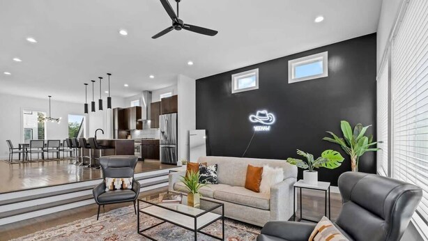 A modern living room with a sofa, coffee table, and chairs, a kitchen in the background, and a black wall with a neon sign.