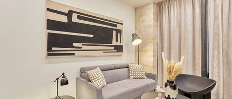 Una sala de estar moderna con un sofá gris, una mesa de centro negra y un sillón negro. En la pared, hay un cuadro abstracto grande, una lámpara de pie y un jarrón con flores secas.