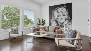 Ein modernes Wohnzimmer mit einer großen Couch, einem Couchtisch und einem Sessel. Große Fenster mit Jalousien, eine Pflanze und ein an der Wand befestigtes Kunstwerk sind ebenfalls zu sehen.