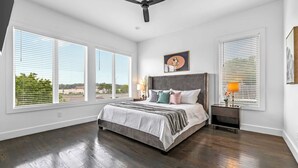 A bedroom with a large bed, two windows with blinds, a ceiling fan, and a nightstand with a lamp.