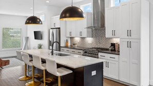 A modern kitchen with white cabinets, a large island with bar stools, a stainless steel range hood, and pendant lighting.