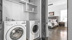A laundry area with two LG Direct Drive washing machines, a bed with a headboard, a bedside table, and a window with blinds.