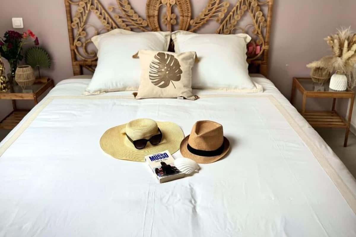 Een slaapkamer met een decoratief hoofdbord, een bed met witte beddengoed, een hoed, zonnebril en een tijdschrift op het bed.