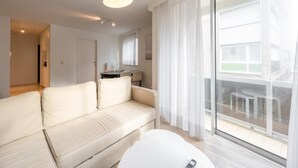 A modern living room with a sofa, a dining table, and a large sliding glass door leading to an outdoor area. - Apartment in Heist Near the Beach (Knokke-Heist)