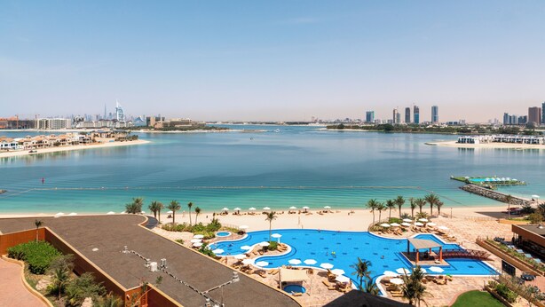 Un resort con una amplia piscina, zona de playa y el perfil urbano al fondo.