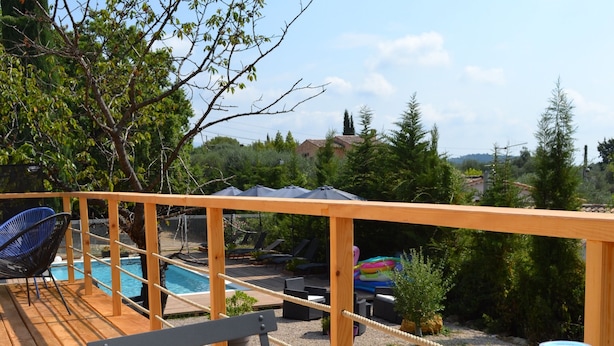 Une terrasse en bois avec une table préparée pour le petit-déjeuner, comprenant des croissants et du jus de fruits, donnant sur une piscine et un espace verdoyant.