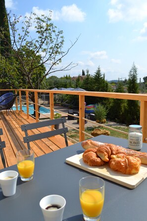 A wooden deck with a table set for breakfast, including croissants and juice, overlooking a pool and greenery. - Le Jardin De Lucette (Le Rouret)