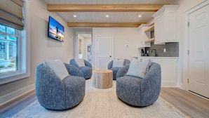 A modern living room with three grey upholstered chairs, a round wooden coffee table, and a large window with a view of a building.