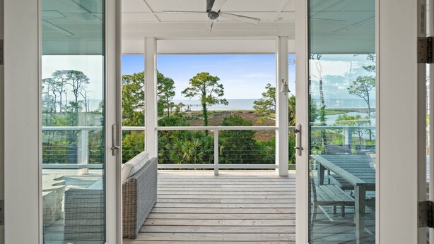 Une vue à travers une porte vitrée donnant sur une terrasse en bois avec un ventilateur de plafond, et offrant un panorama sur un paysage arboré et un plan d’eau.
