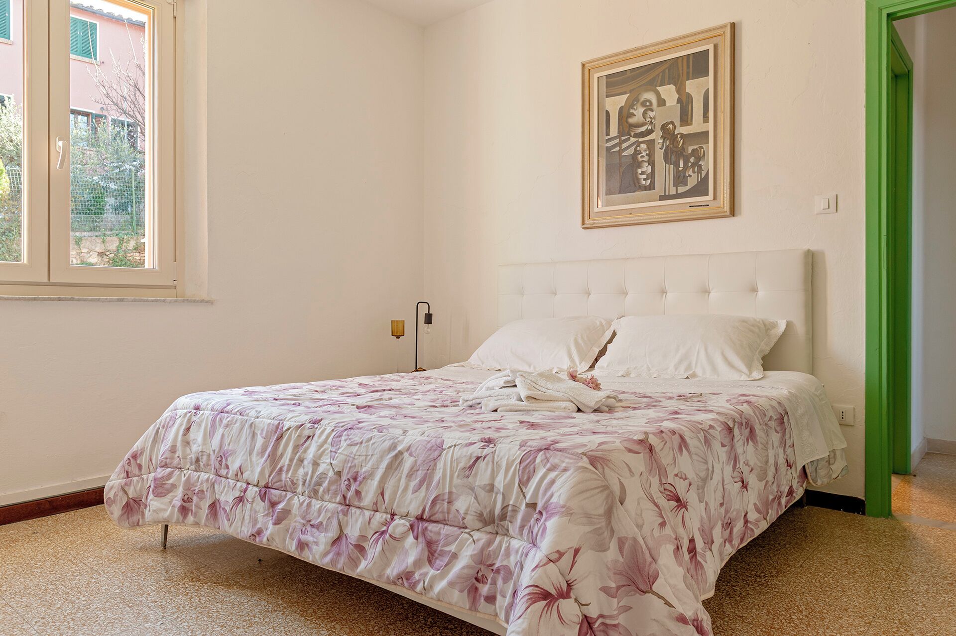 A bedroom with a bed, a painting on the wall, and a window with a view of buildings.