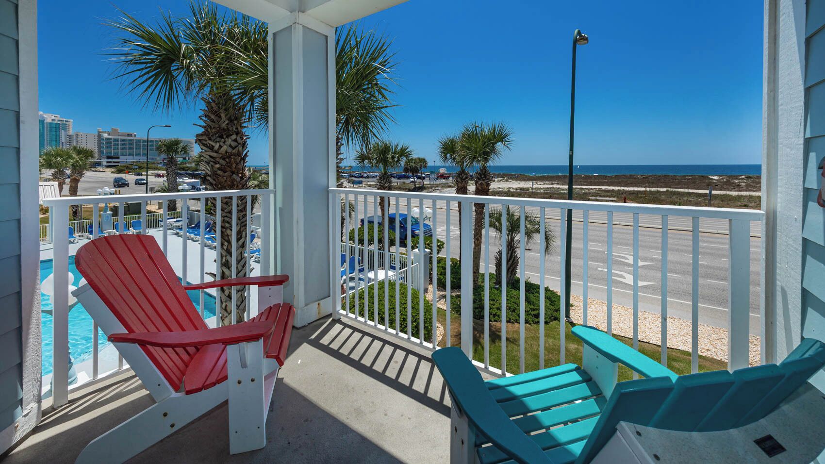 A balcony with two red and two blue Adirondack chairs, a pool, and a view of the beach and ocean.