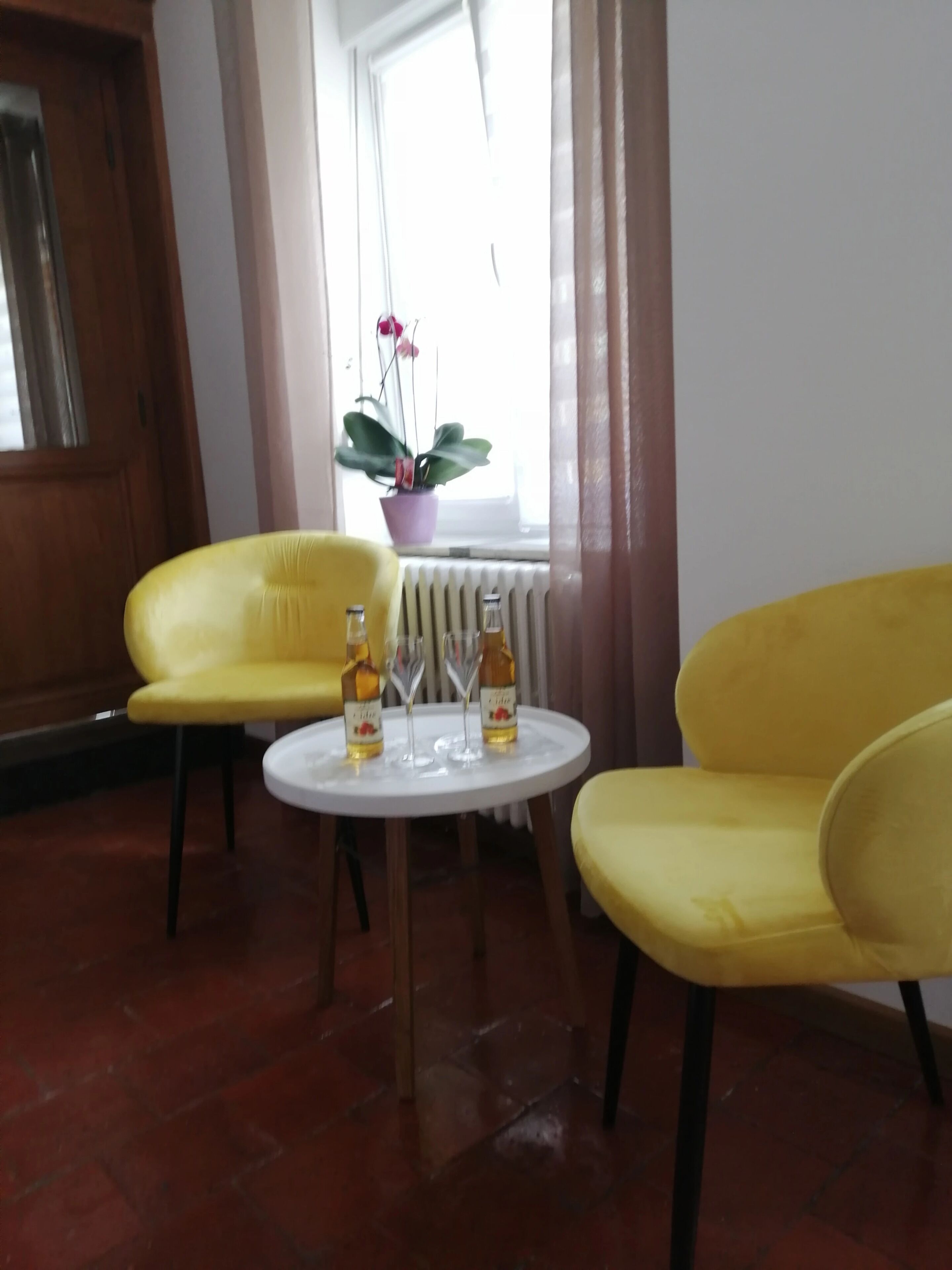 Una habitación con dos sillas amarillas, una pequeña mesa blanca con botellas y vasos, una planta en maceta y una ventana con cortinas.