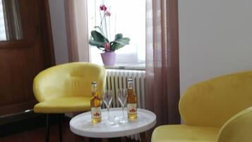 Una habitación con dos sillas amarillas, una pequeña mesa blanca con botellas y vasos, una planta en maceta y una ventana con cortinas.