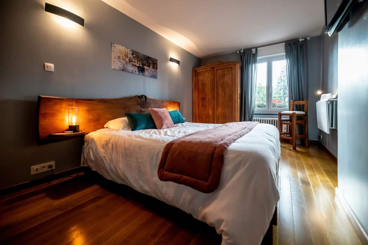 Une chambre à coucher avec un grand lit, une tête de lit en bois, des lampes de chevet, un tableau au mur, une fenêtre avec des rideaux et un radiateur.