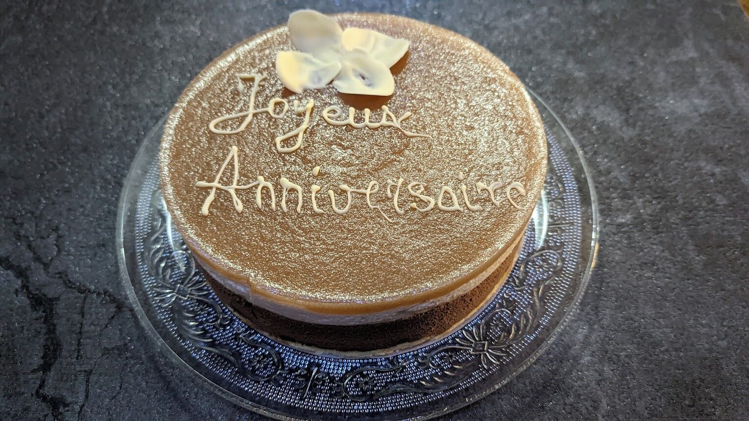A round cake with "Joyeux Anniversaire" written on top, placed on a decorative glass plate.