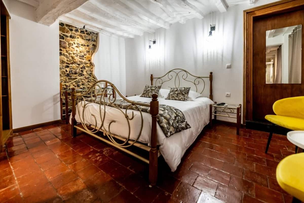 Une chambre avec un mur en pierre, un lit avec un cadre en métal, une table de chevet et une chaise jaune.
