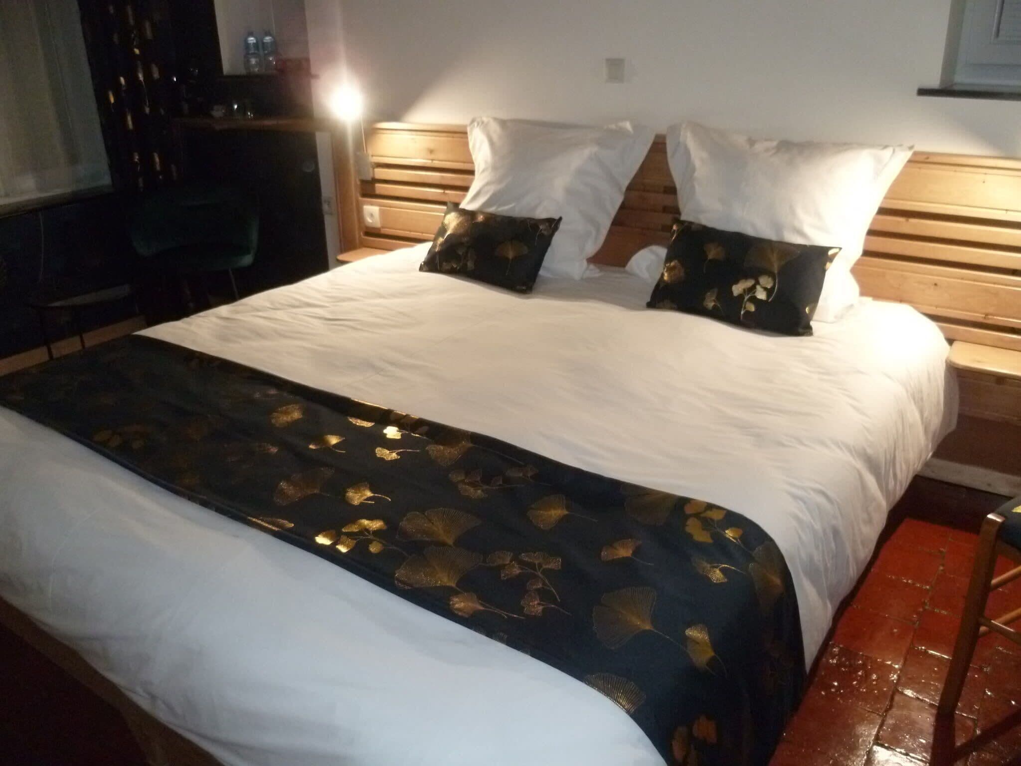 A hotel room with a large bed, two pillows, a wooden headboard, and a dark blue bedspread with gold leaf patterns.