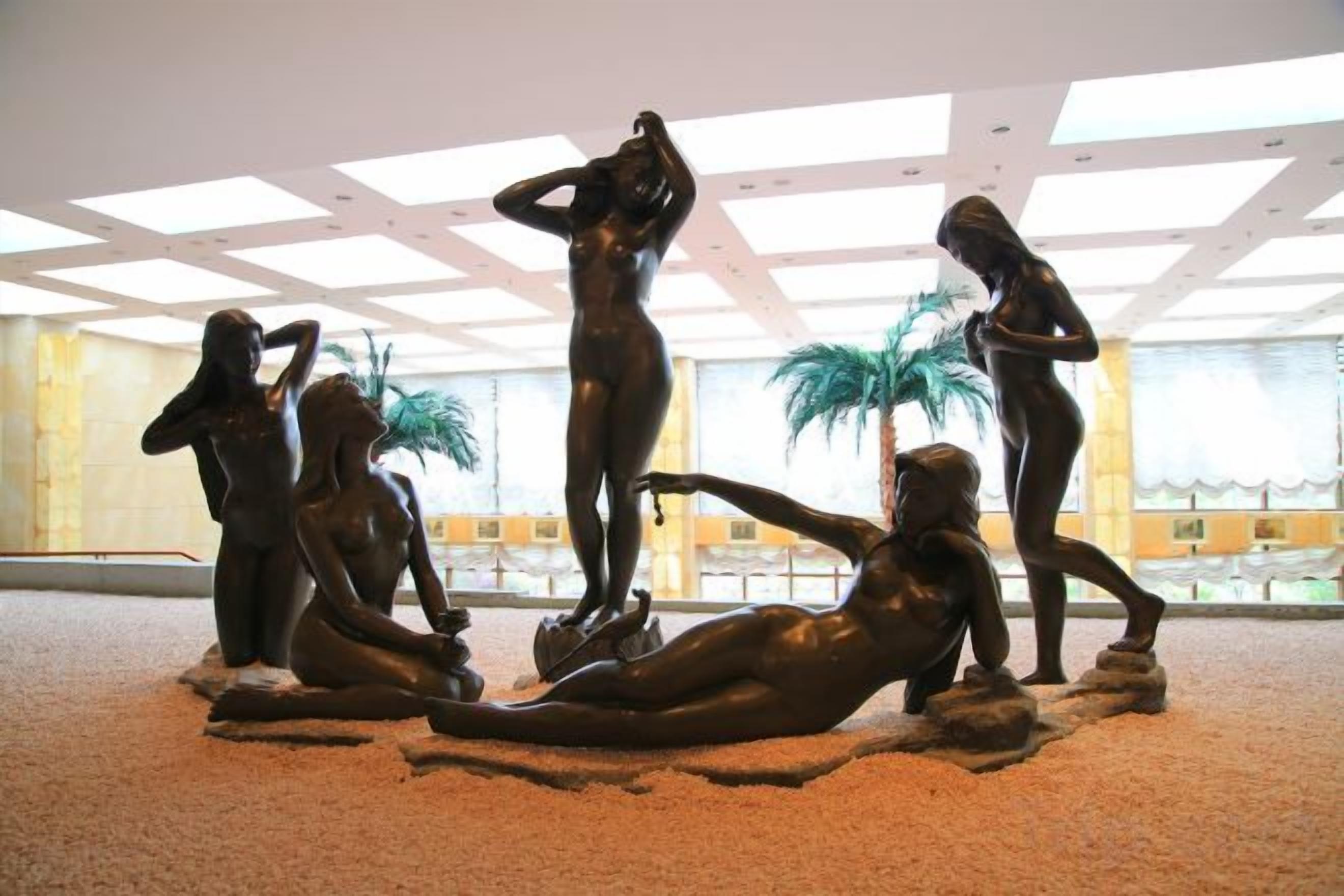 A sculpture of human figures in various poses, set on a textured floor, in a room with a high ceiling and large windows.