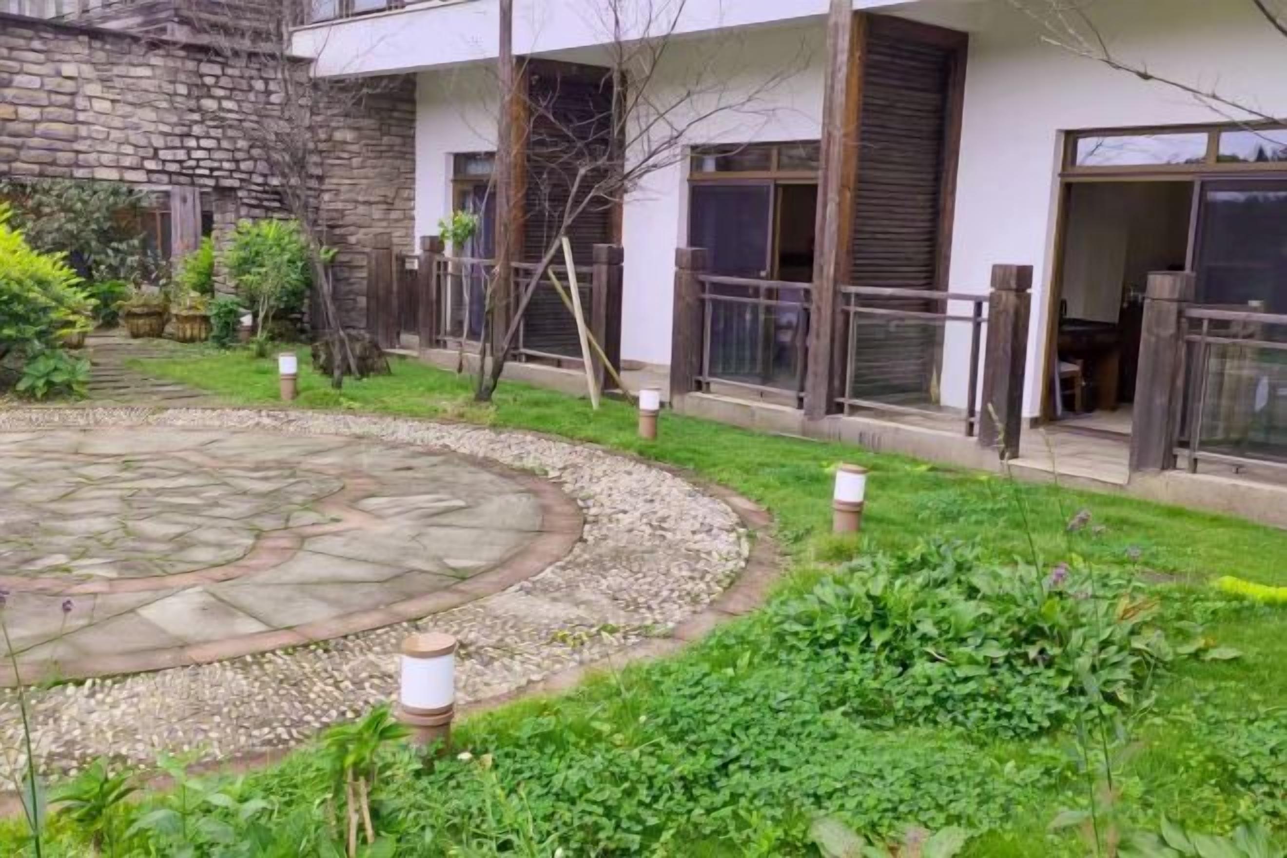 Un pati amb un paviment de pedra circular envoltat de grava, una zona verda amb plantes, i un edifici amb façana blanca i grans finestres.