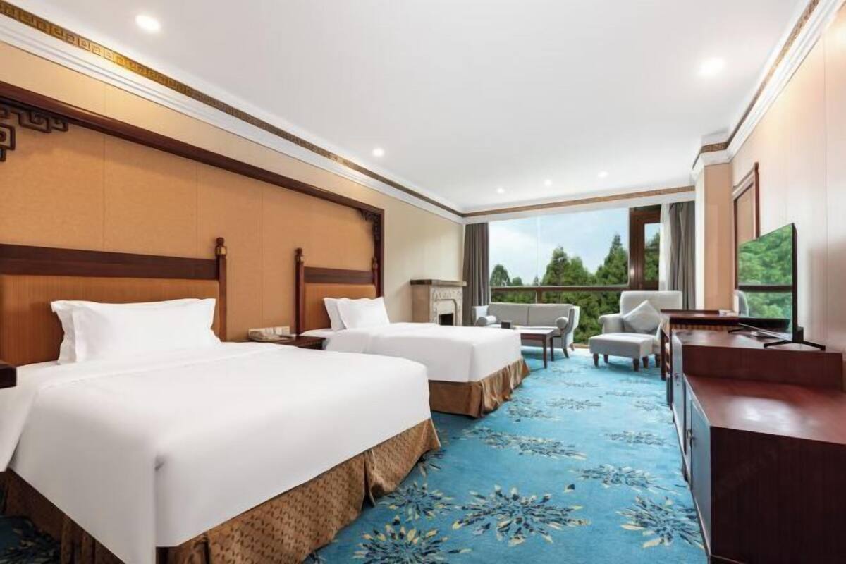 A hotel room with two beds, a desk, a chair, and a large window with a view of greenery.