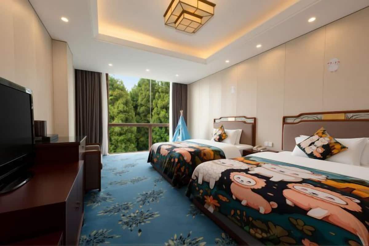 A hotel room with a TV, two beds with colorful bedding, a large window with a view of trees, and a ceiling light fixture.