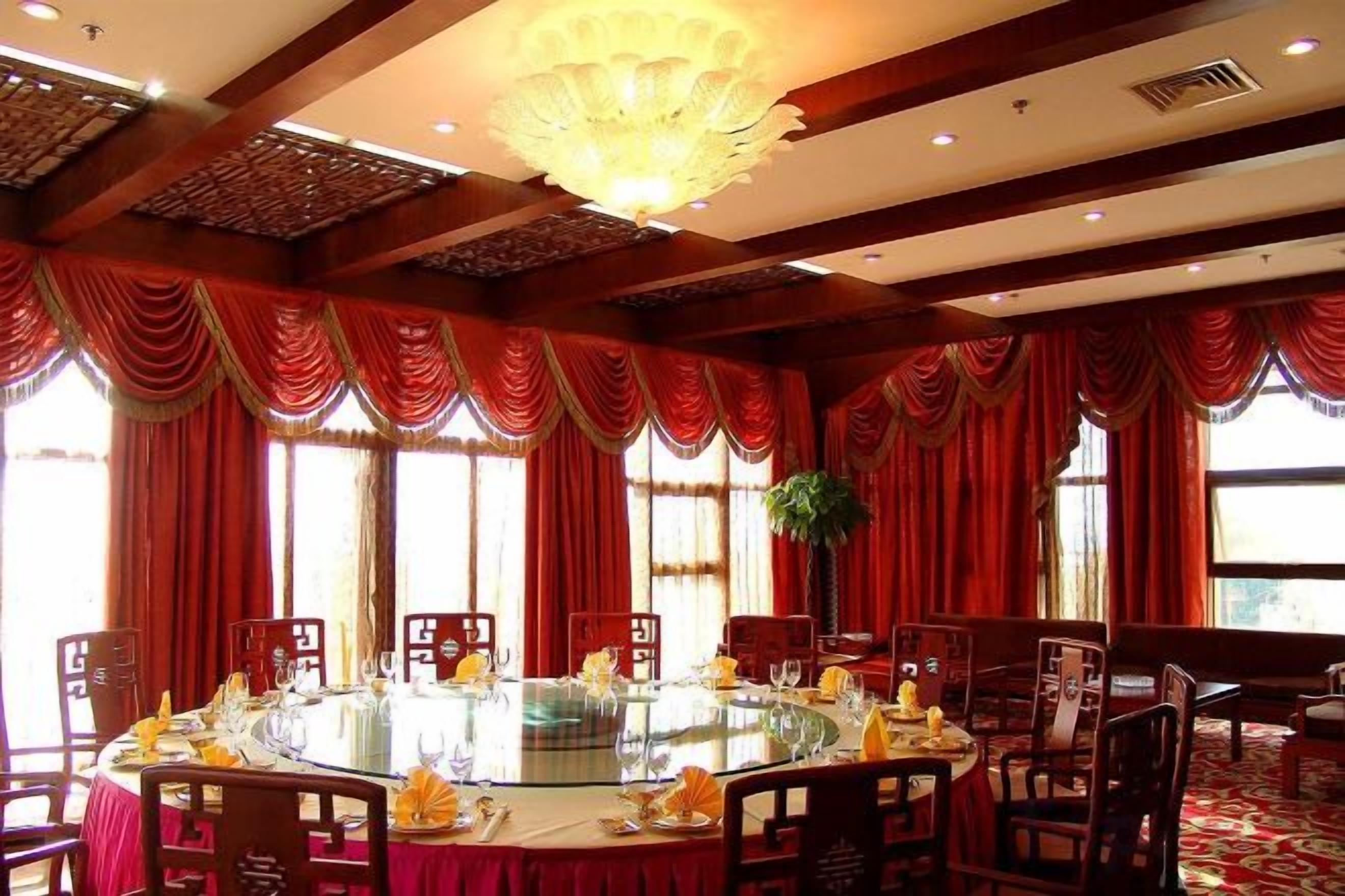 A round table with chairs, red curtains, and a chandelier in a room with wooden beams.