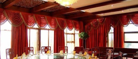 A round table with chairs, red curtains, and a chandelier in a room with wooden beams.