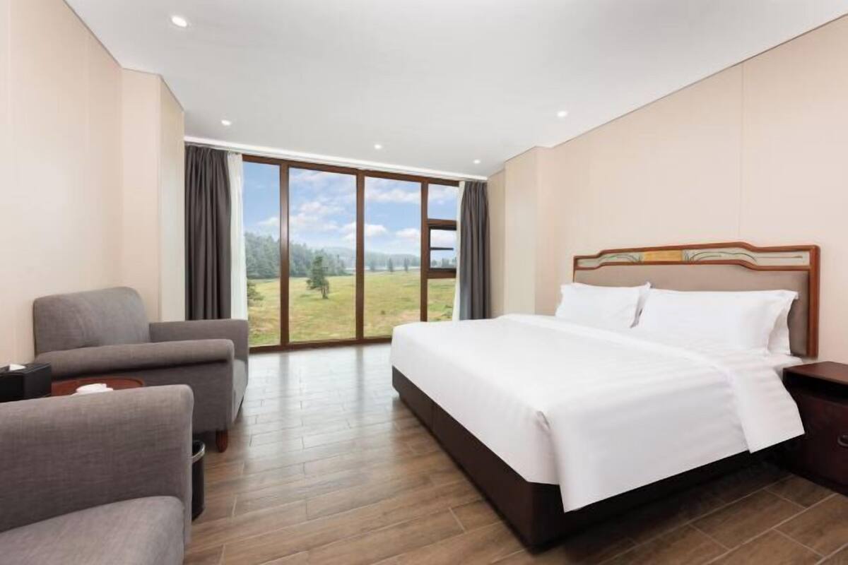 A hotel room with a large bed, a sofa, a nightstand, and a view of a grassy field through a large window.
