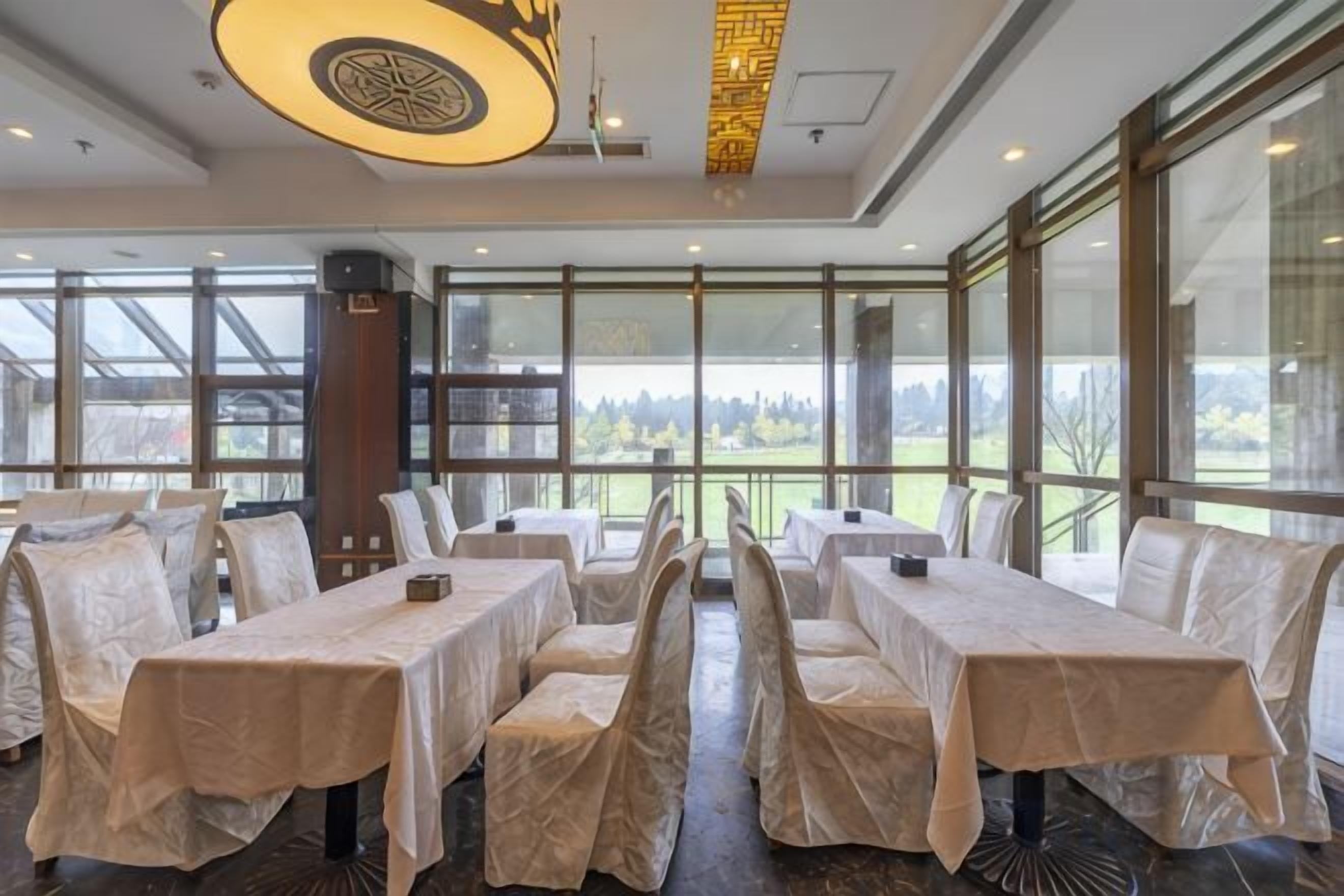 Restaurante com mesas cobertas por toalhas brancas, grandes janelas e vista para o exterior.