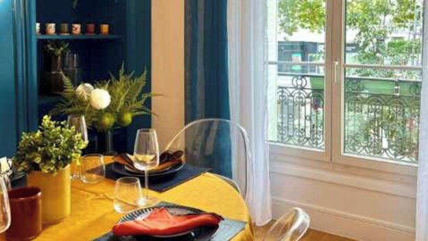 A dining area with a table set for two, a bookshelf with decorative items, and a view of the outside through a large window. - Elegant Apartement - AC 2br-6p- Palais des Congres (Paris)